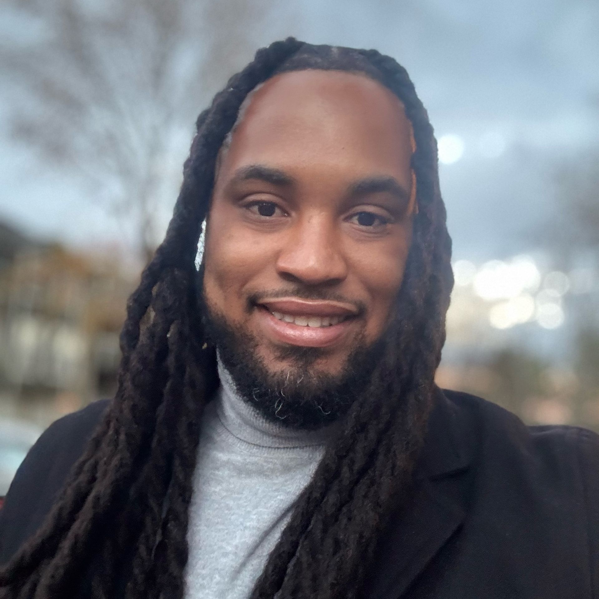 Man with long dreadlocks, gray turtleneck, black jacket smiles outdoors.