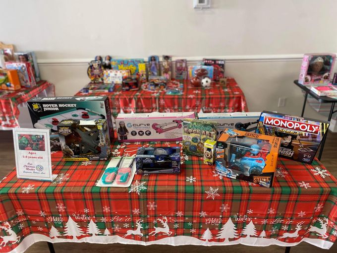 Tables covered in red plaid tablecloths, laden with wrapped presents and toys, set up for a holiday event.