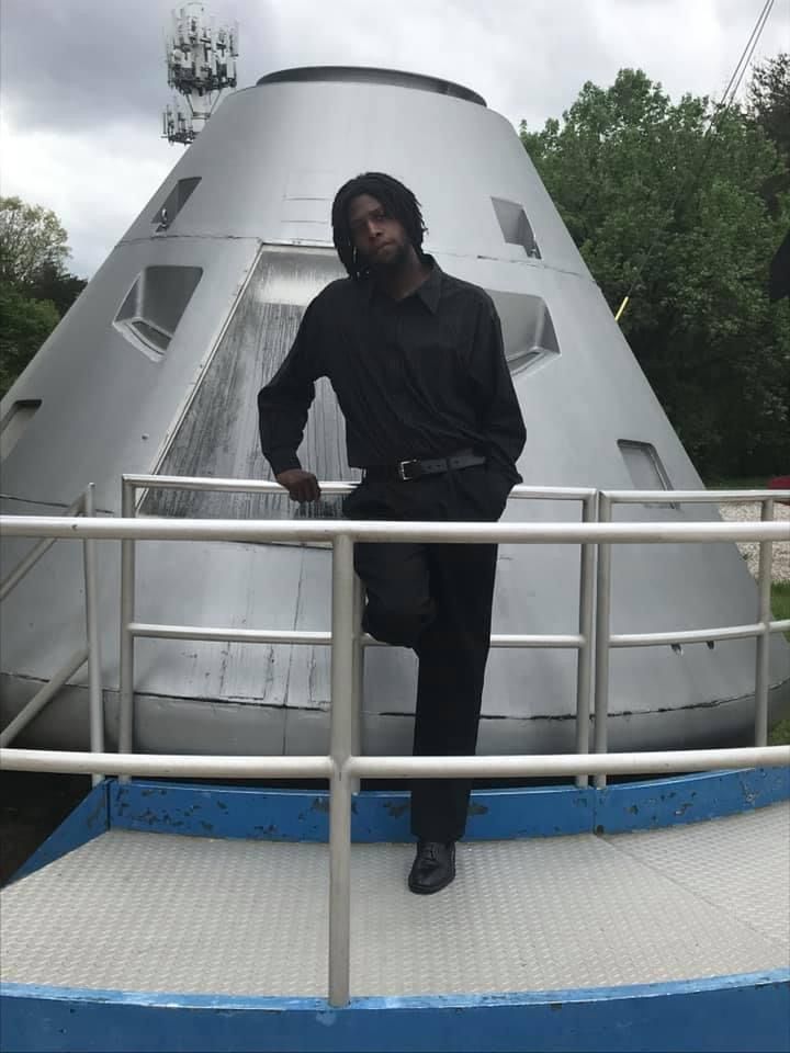 A man in a black shirt leans on a railing in front of a cone shaped building
