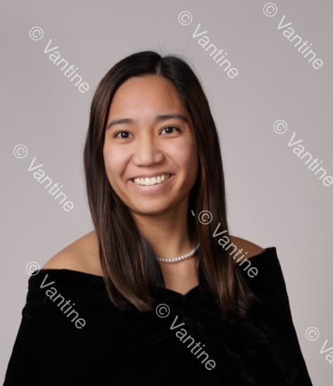 Woman with a pearl necklace, smiling, wearing a black off-the-shoulder top, against a grey background.