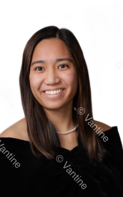 Woman with long brown hair, wearing a black off-the-shoulder top and pearl necklace, smiling against a white backdrop.