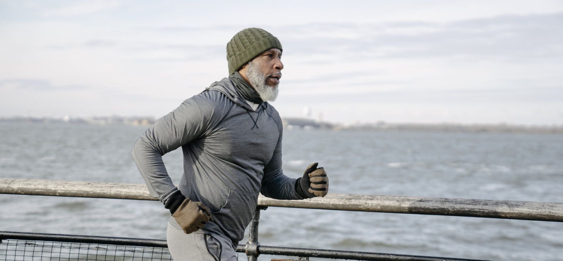 Man runs along waterfront wearing a beanie, gloves, and grey athletic gear.