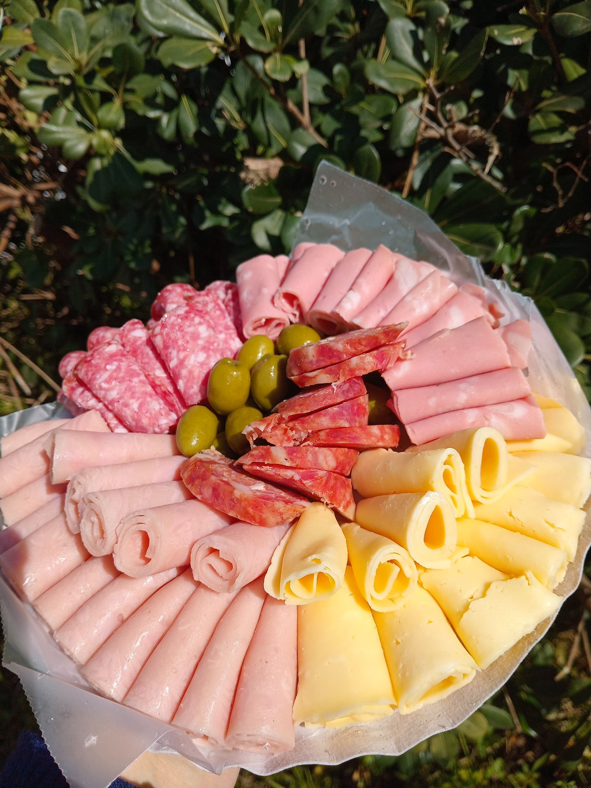 Plato de embutidos con salami, jamón, queso, aceitunas y salami, dispuestos sobre una superficie blanca, al aire libre.