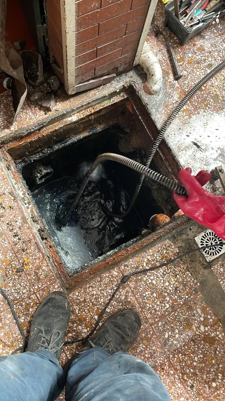 Escotilla de servicio abierta con una manguera y guantes rojos junto a una pared de ladrillos y un suelo mojado y salpicado.