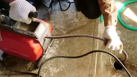 Un trabajador con guantes utiliza una máquina roja con mangueras para limpiar un suelo mojado y un desagüe.