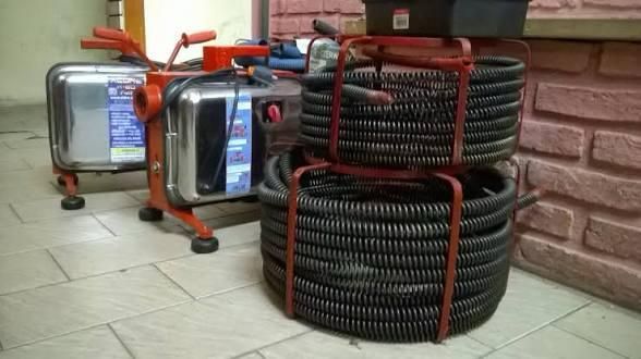 Mangueras negras enrolladas sujetas con correas rojas junto a unidades de climatización plateadas sobre un suelo de baldosas.