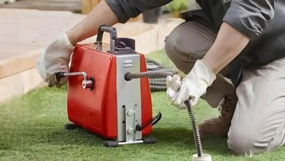 Un técnico se arrodilla junto a una máquina roja desatascadora sobre el césped, manipulando una manguera metálica enrollada.