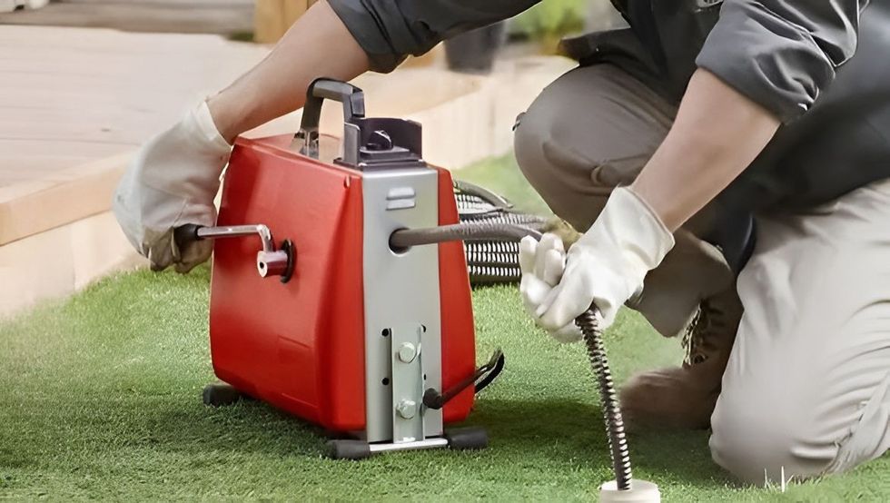 Una persona arrodillada sobre la hierba, junto a una máquina portátil roja, ajusta un cable con las manos enguantadas.