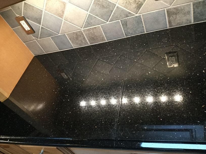 Black speckled countertop with backsplash, inset lighting, and grey-brown tile.