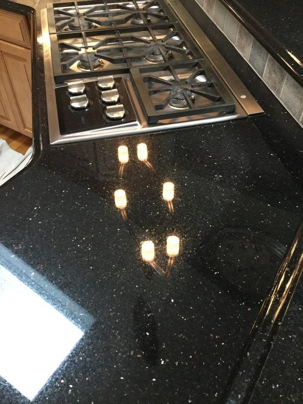 Black granite countertop with the reflection of six light bulbs. A stainless steel gas stovetop sits behind the countertop.