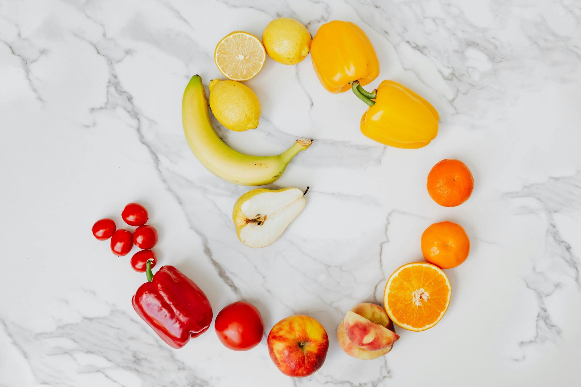 Fruits and vegetables arranged in a colorful circle on a marble surface: yellow peppers, lemons, oranges, red pepper, tomatoes, and other fruits.