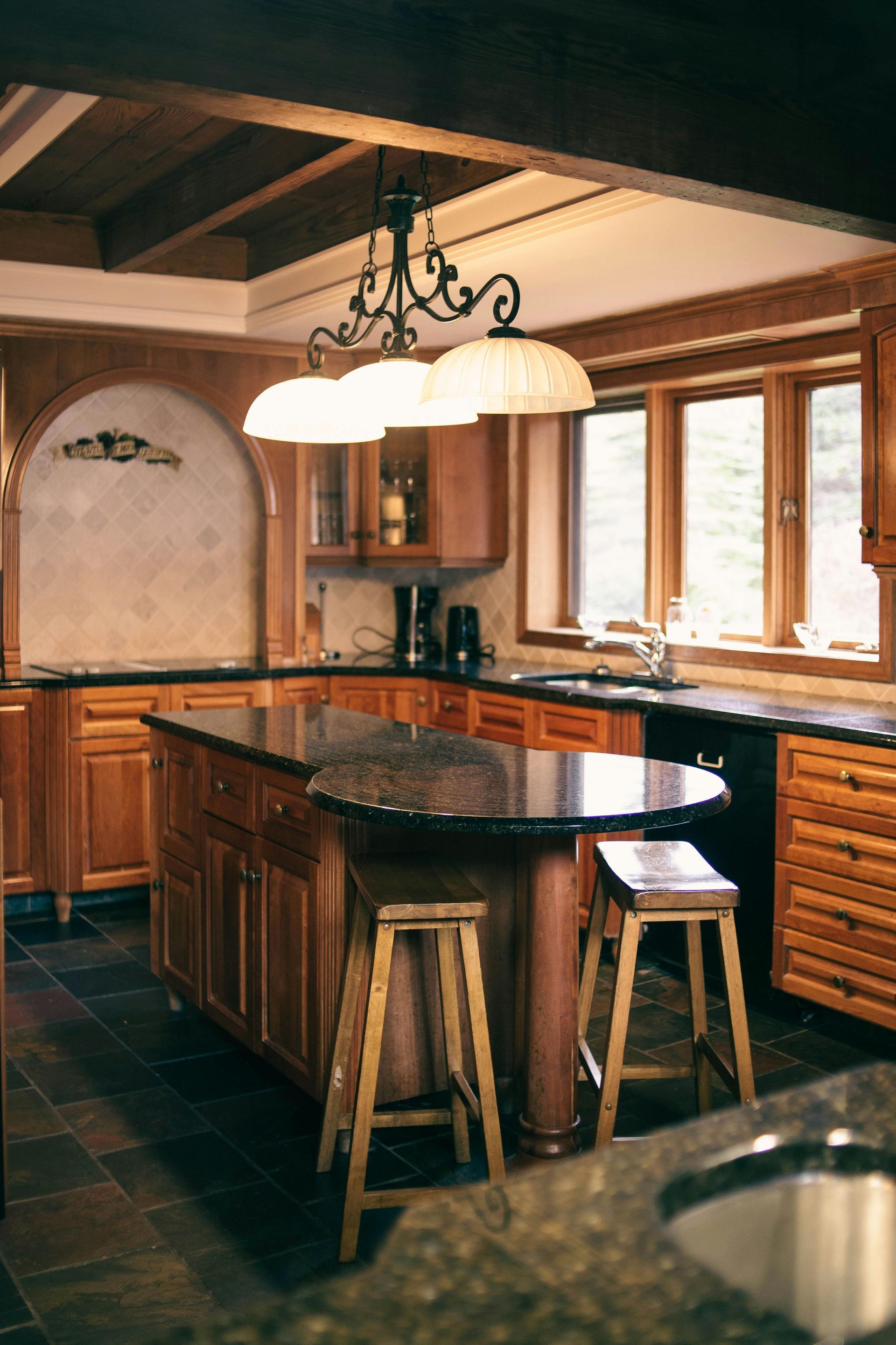 Kitchen with wooden cabinets, a dark countertop, and an island with stools; overhead chandelier and windows.