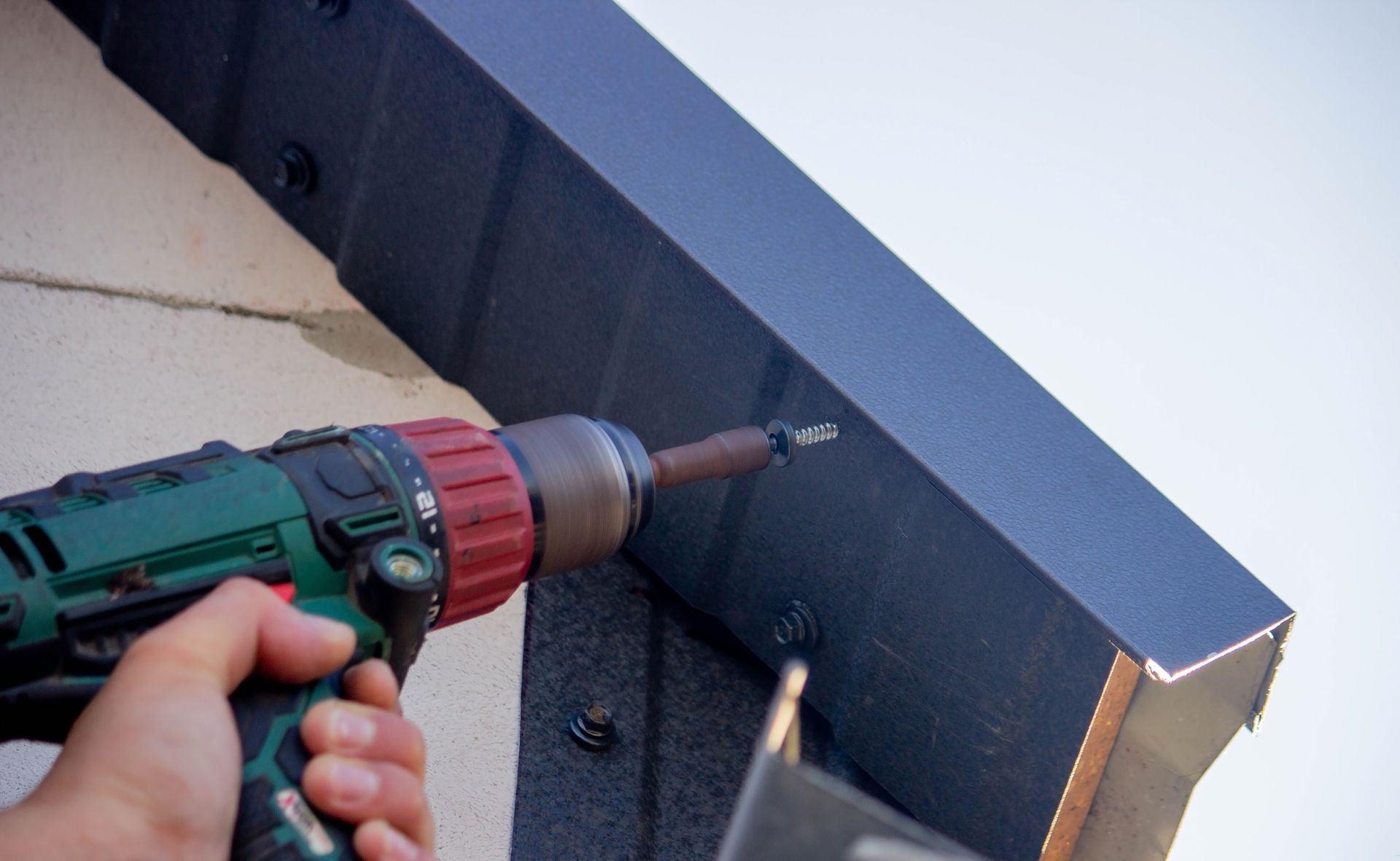 A hand uses a green power drill to drive a screw into a dark gray metal trim piece installed on a wall.
