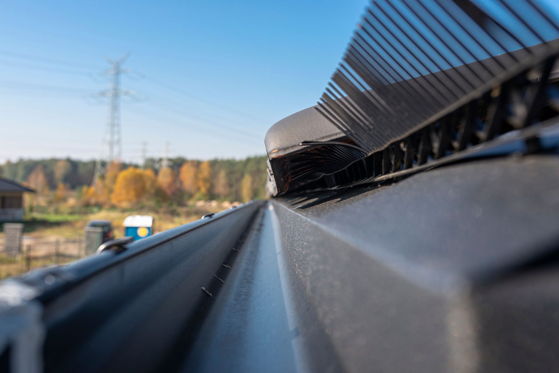 A close-up, low-angle view of a dark metal roof gutter featuring a black plastic leaf guard against a clear blue sky.