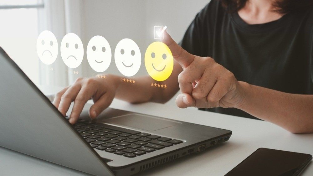 A person using a laptop clicks a yellow smiley face icon on a screen to indicate a five-star positive customer rating.