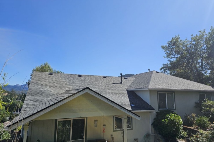 A tan house with a gray shingle roof, set against a backdrop of trees and hills on a sunny day.