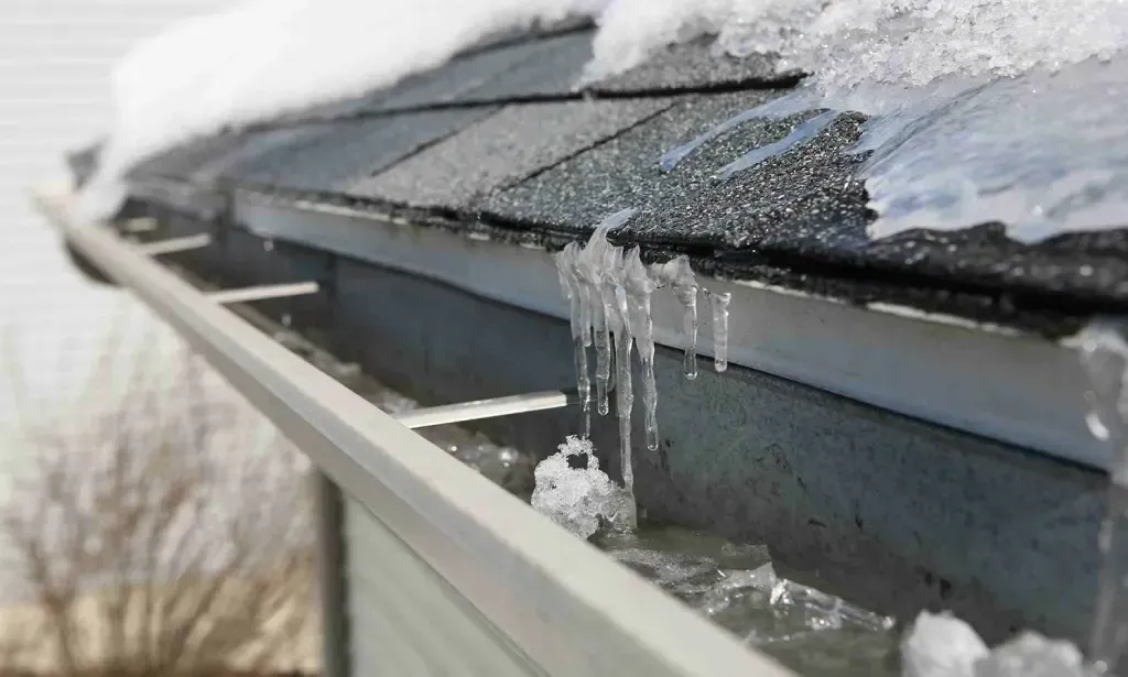 Close-up of a house gutter filled with ice and snow, with icicles hanging from the edge of the roof shingles.