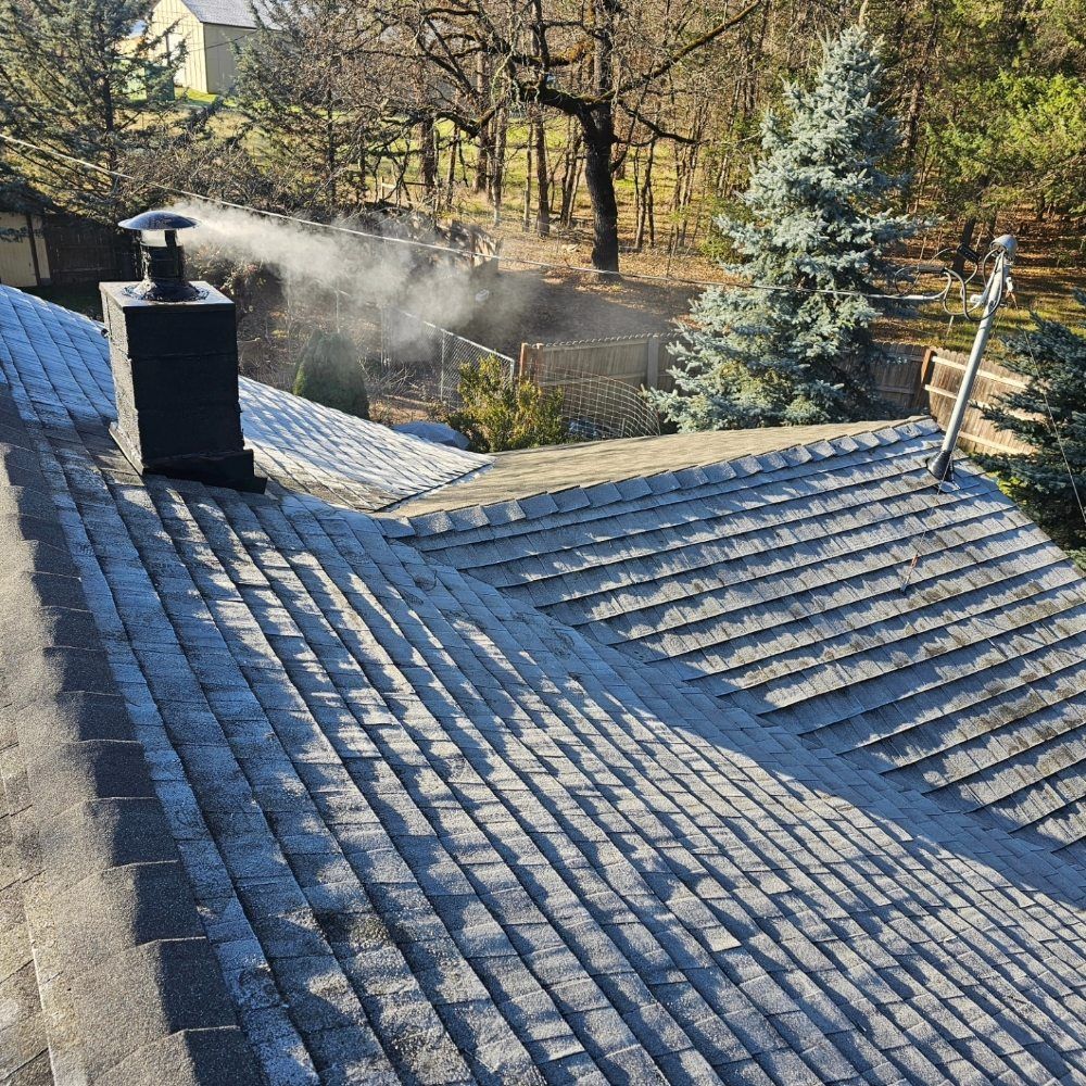 A shingled roof with a black chimney emitting smoke on a sunny day with trees in the background.