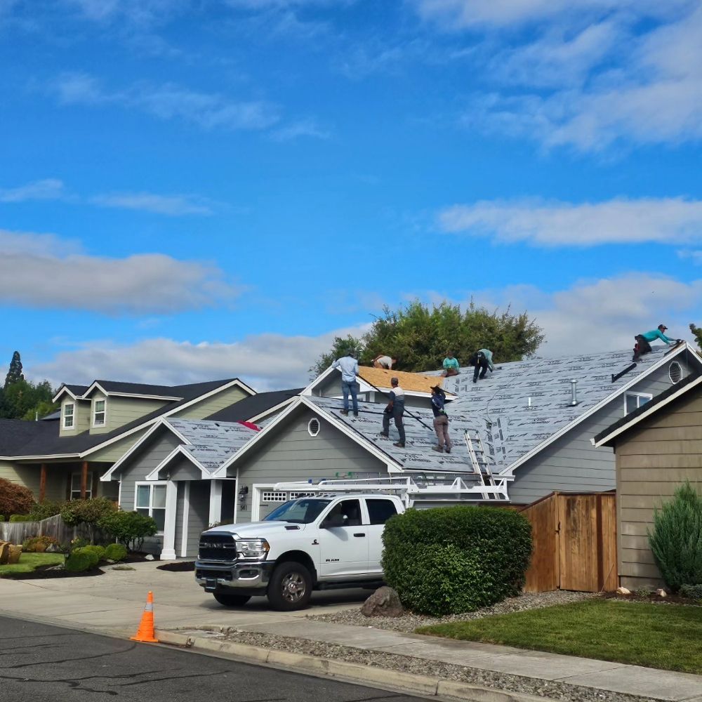 Workers install new roofing on a light-colored suburban house with a white truck parked in the driveway.