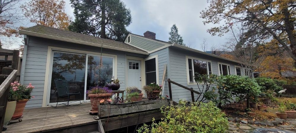 A blue, single-story house with a wooden deck, sliding glass door, and surrounding trees during the autumn season.