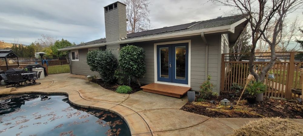 A grey house with blue French doors opens onto a backyard patio next to a swimming pool under a cloudy sky.