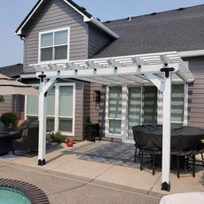 White pergola attached to a gray house with a patio, outdoor furniture, and a pool in the foreground.