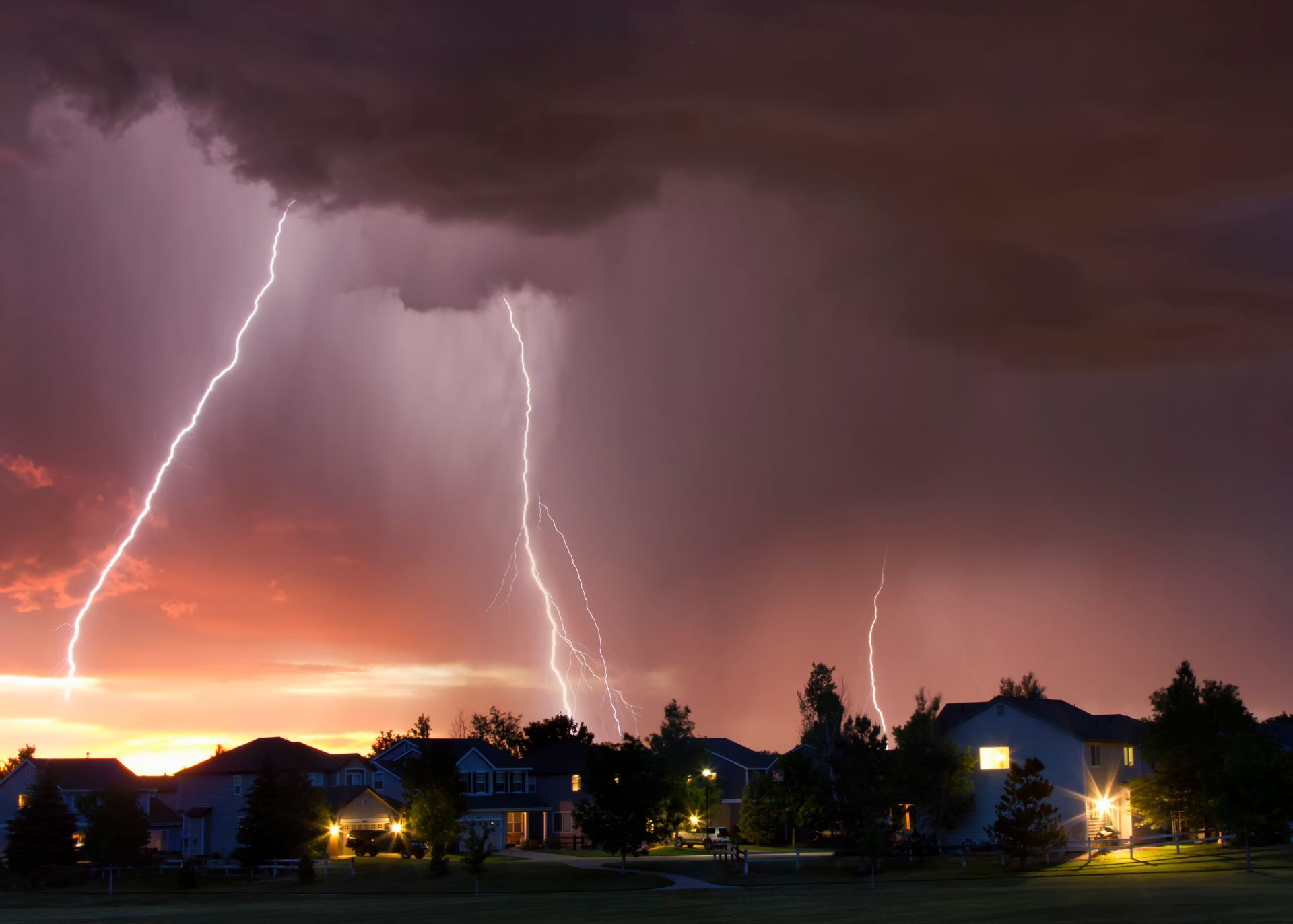 Multiple lightning bolts strike behind a row of suburban houses during a sunset storm.