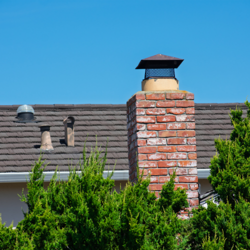 Brick chimney on a house roof, blue sky, surrounded by green bushes.