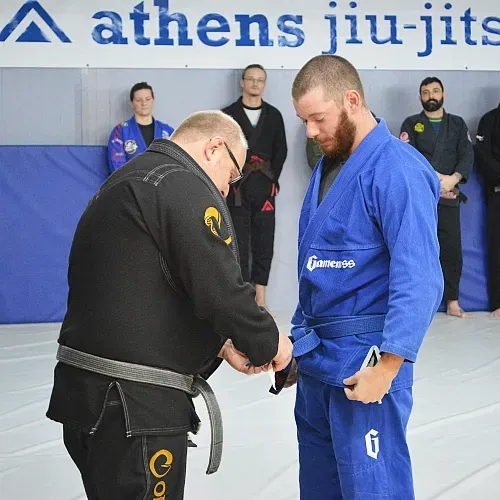 Man in blue gi receiving belt from instructor in black gi at Athens Jiu-Jitsu.