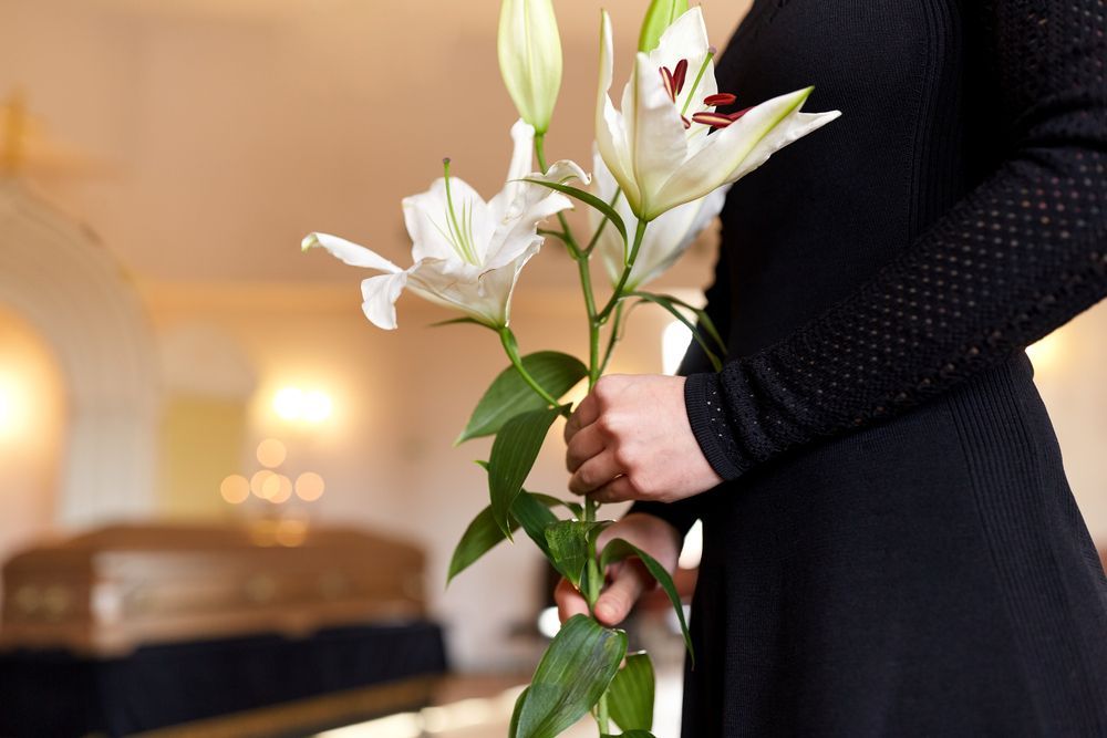 A person in black clothing holds white lilies in front of a coffin at a funeral service — Mid Coast Funeral and Cremation Service In Port Macquarie, NSW