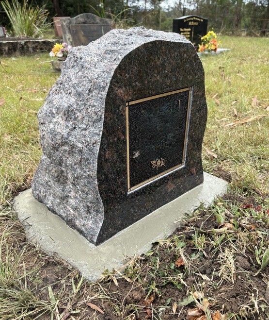 A dark polished granite headstone with a rough-hewn edge sits on a concrete base in a grassy cemetery — Mid Coast Funeral and Cremation Service In Port Macquarie, NSW