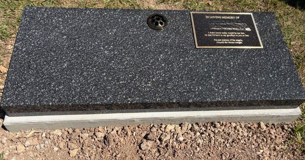 A rectangular granite headstone with a bronze memorial plaque sits on a concrete base in a grassy area.