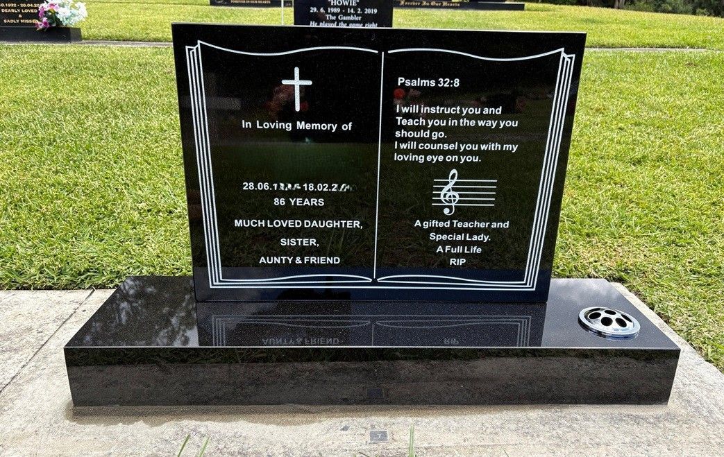 A black granite tombstone shaped like an open book, with inscribed text, a cross, and a musical staff, set in a graveyard — Mid Coast Funeral and Cremation Service In Port Macquarie, NSW