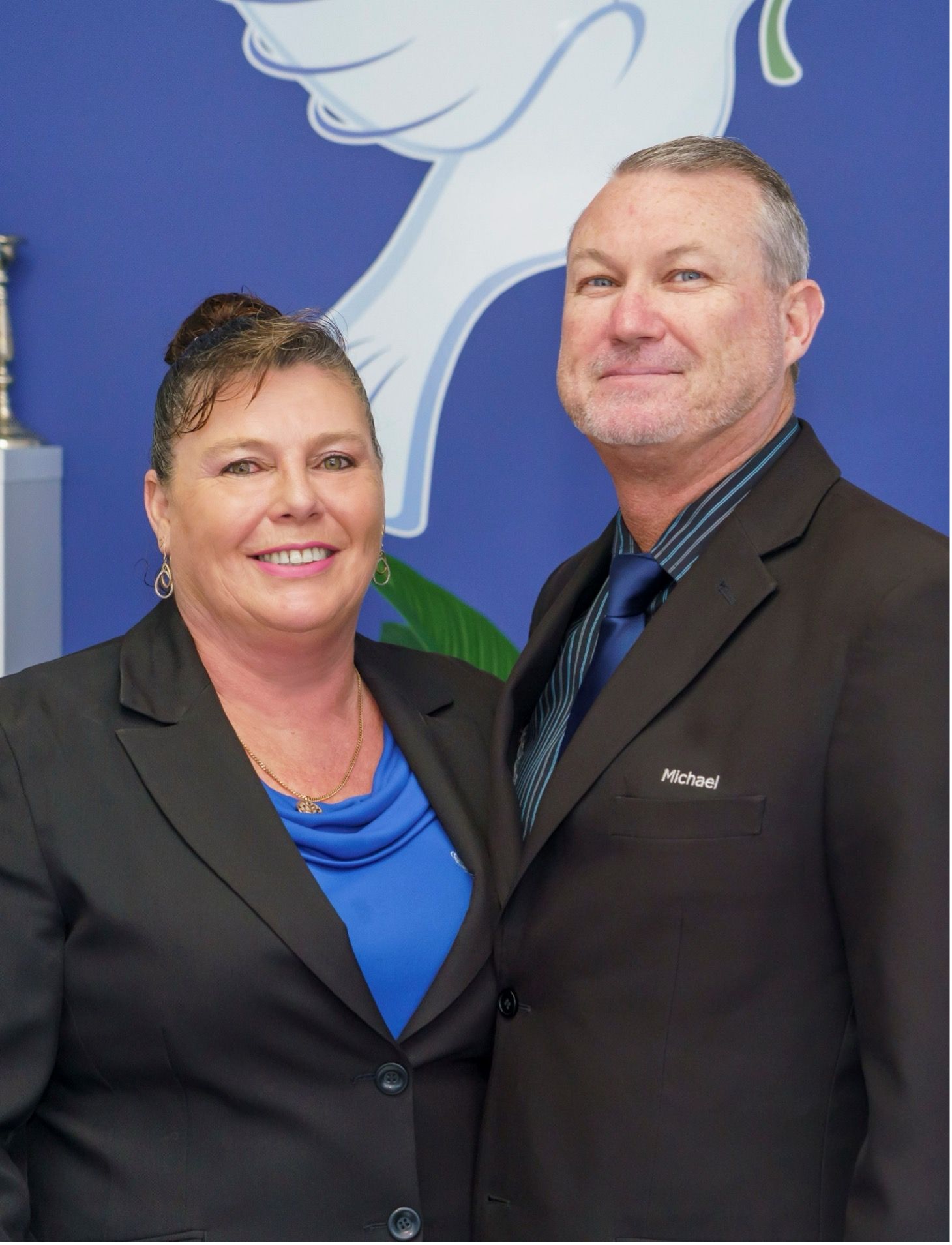 A smiling man and woman wearing professional business suits stand together against a blue background with a white graphic.