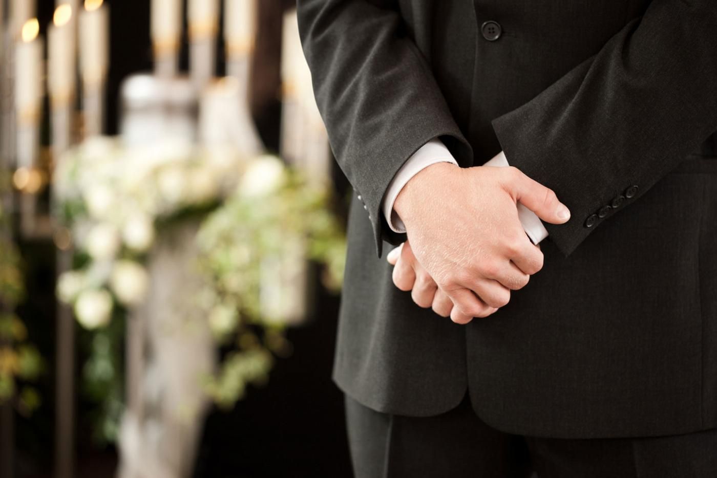A Man in a Suit is Standing With His Hands Folded — Mid Coast Funeral and Cremation Service In Port Macquarie, NSW