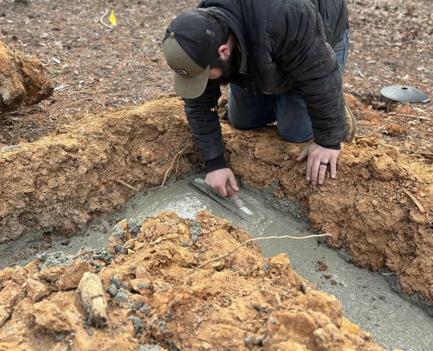 Person kneeling, smoothing concrete in a trench with a trowel outdoors.