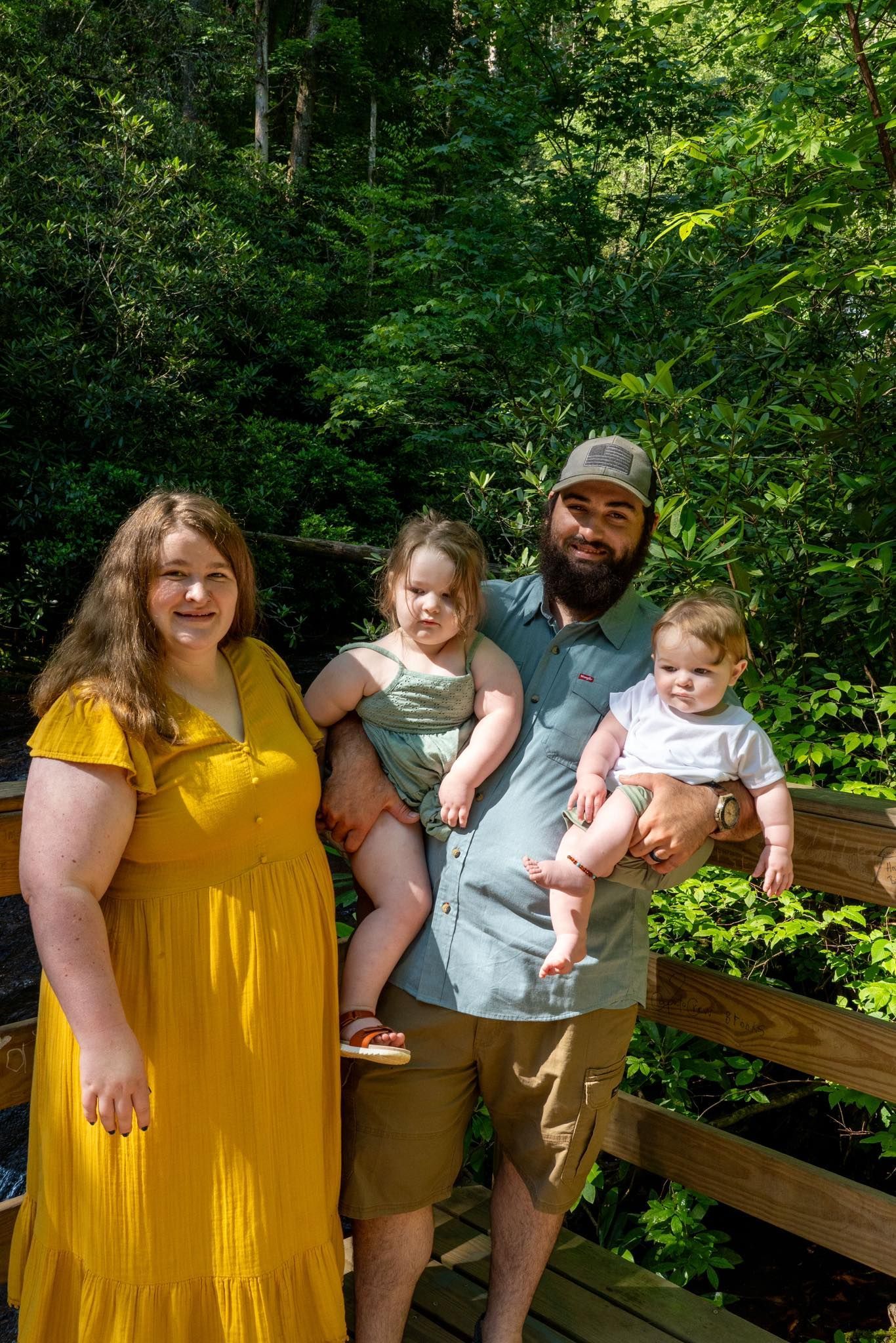 Family of four smiles outdoors: woman in yellow dress, man holding a baby, child in green, surrounded by lush green trees.