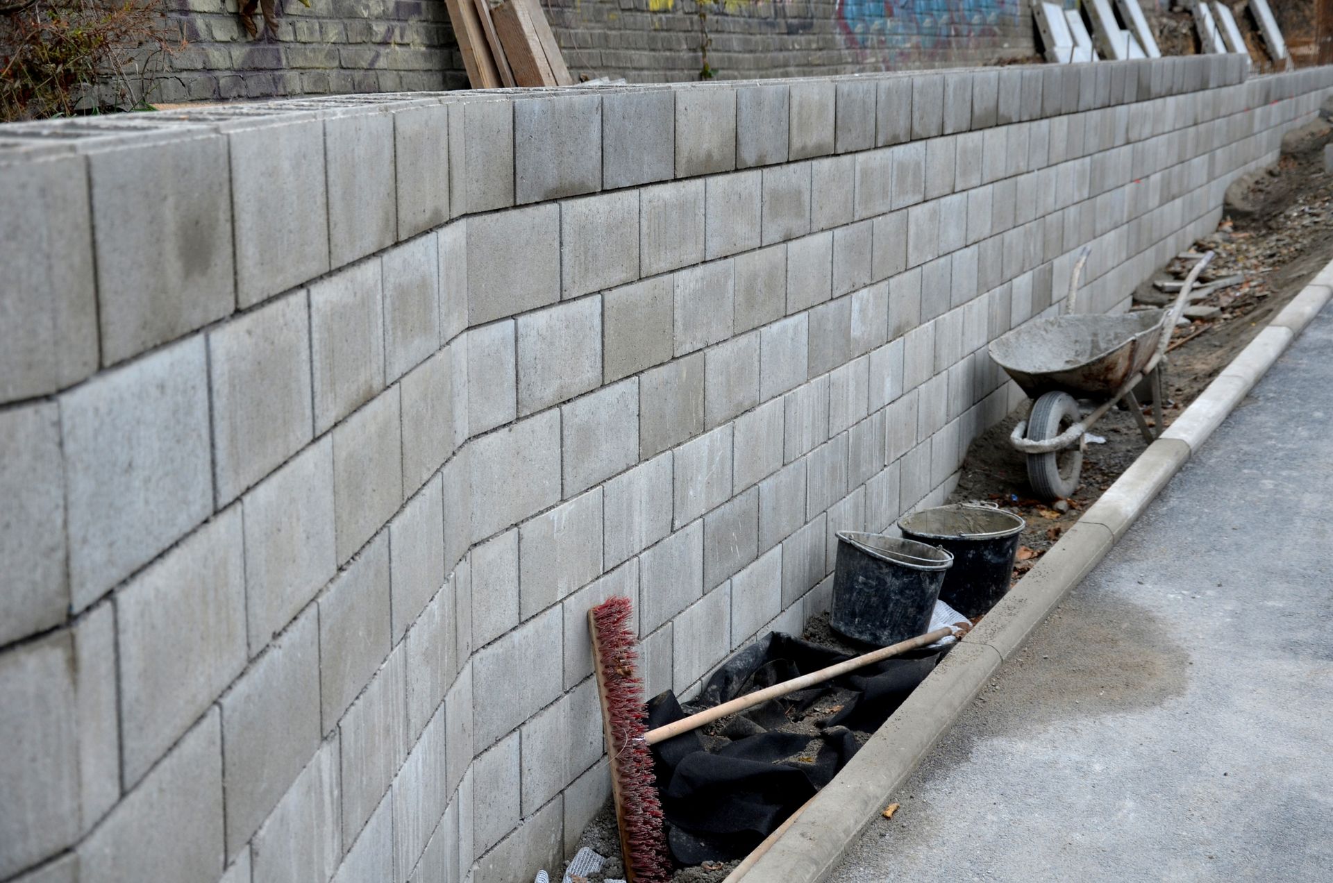 Retaining wall made of gray concrete blocks. Construction materials and wheelbarrow nearby.
