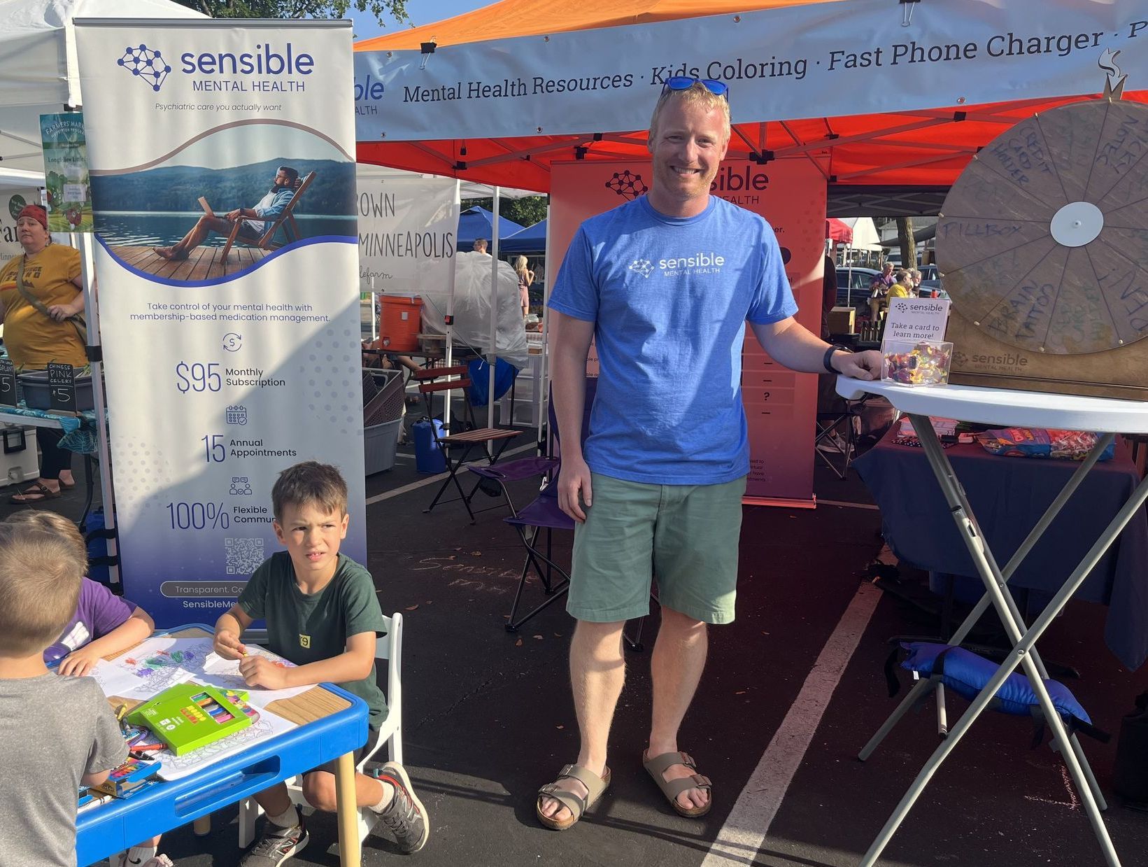 Man at booth with product sample, kids coloring. Outdoor market, sunny day.