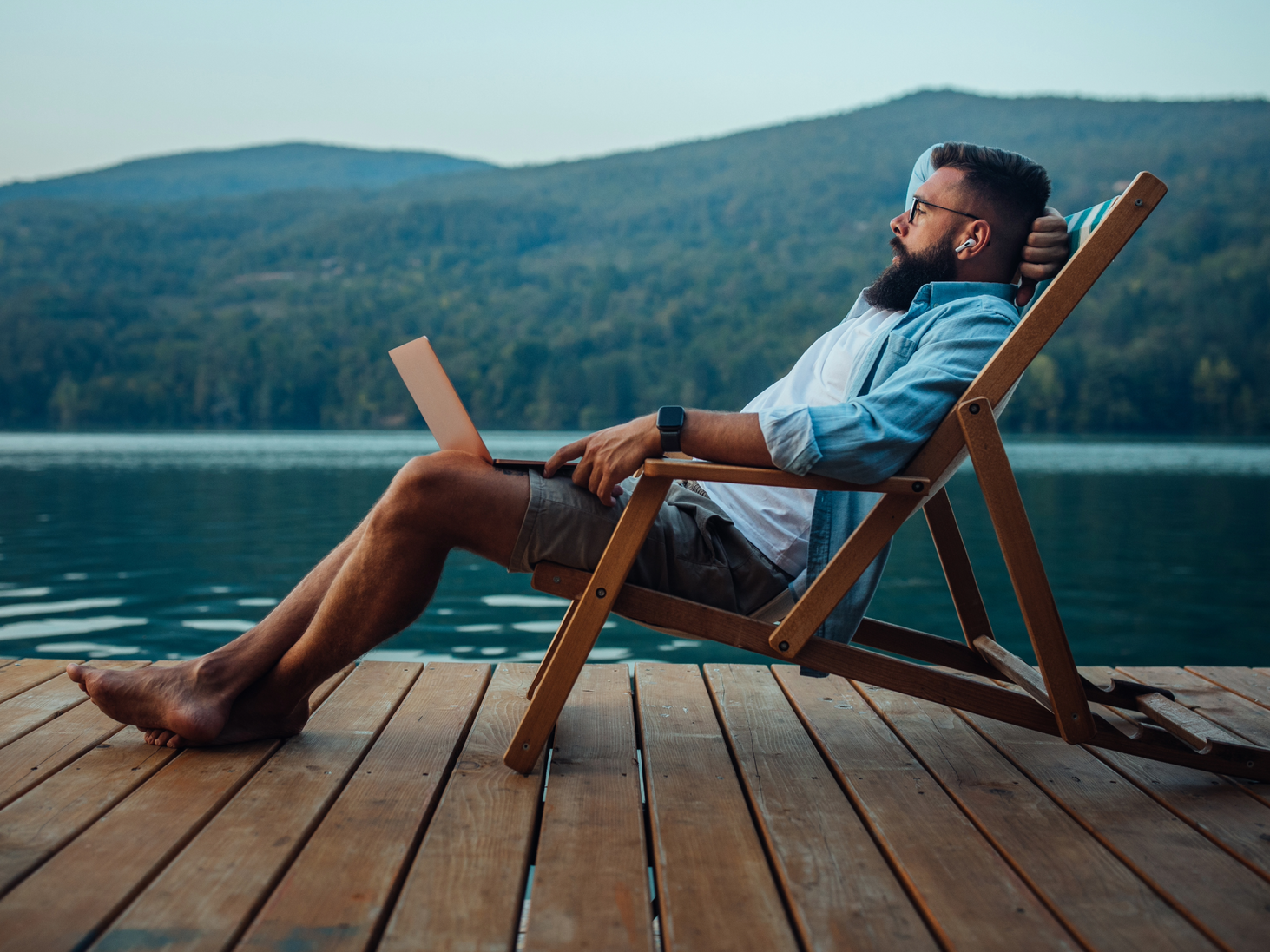 Man in casual clothes, relaxing on a dock with laptop, overlooking a lake and mountains.