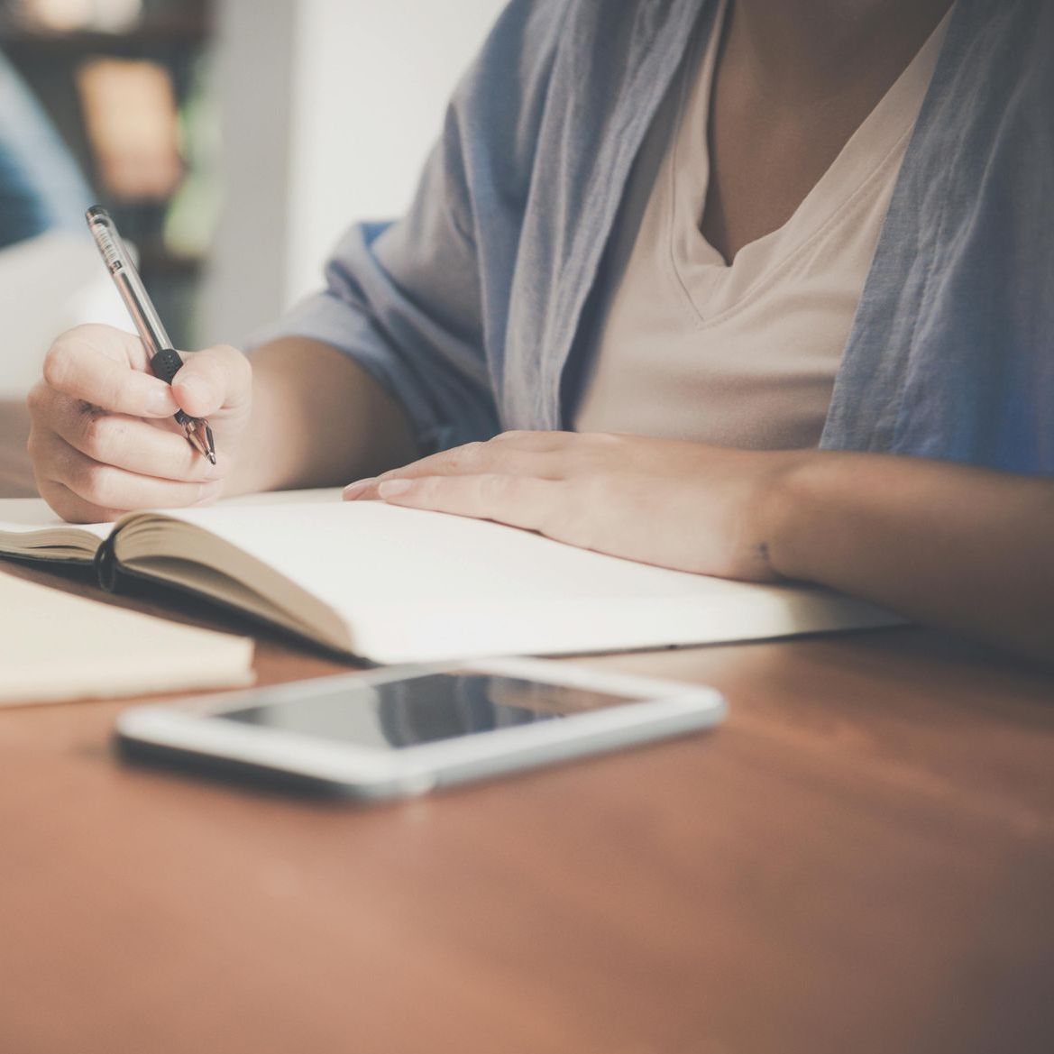 Person writing in a notebook with a pen, a smartphone on the table.