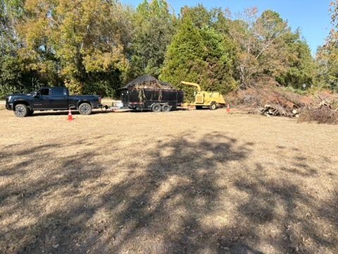 Black truck towing a trailer with mulch, wood chipper, and pile of branches in a field.