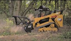 Yellow Caterpillar skid steer with brush cutter in a wooded area.