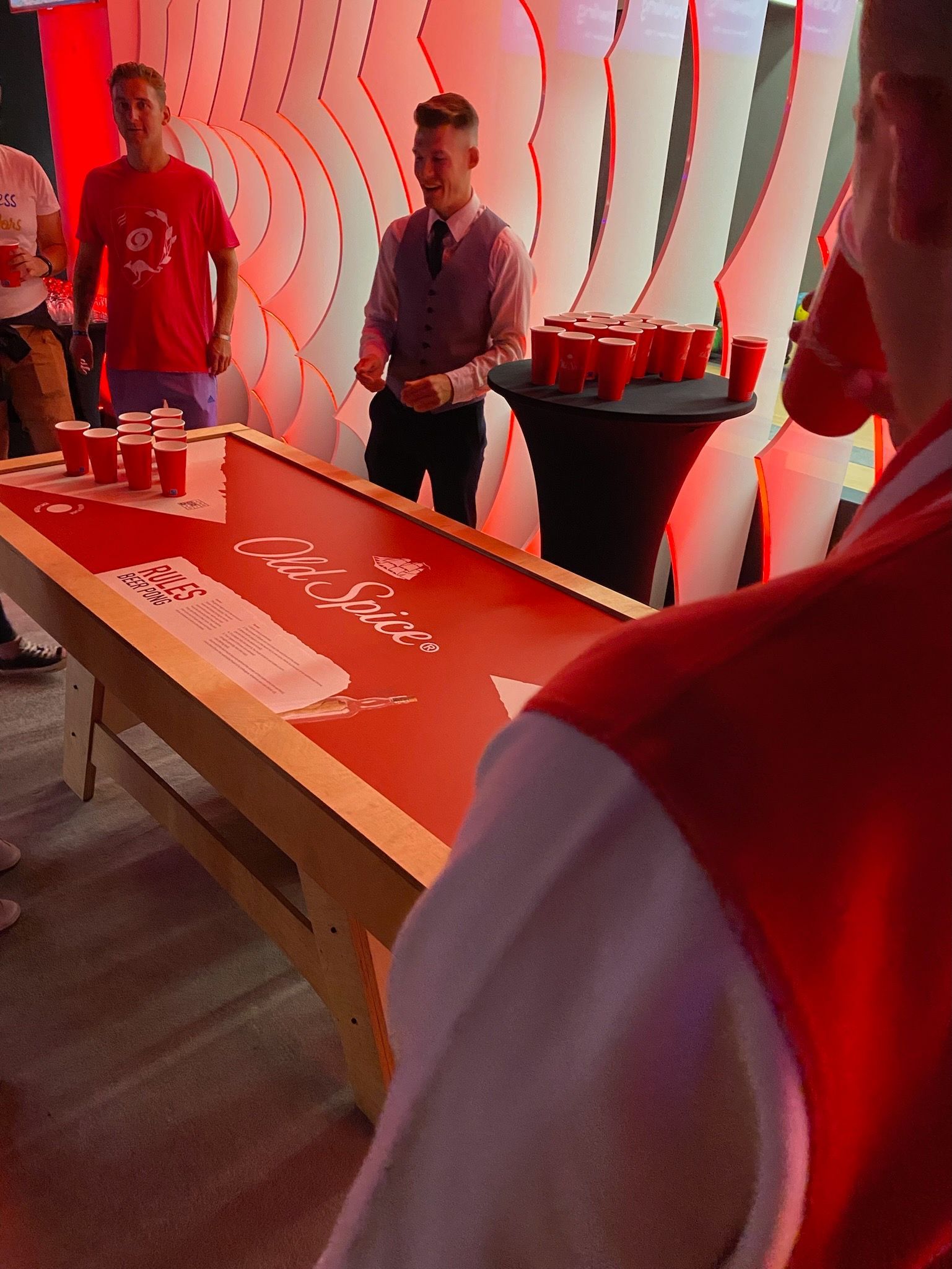 mężczyzna w czerwonej koszuli stoi przed stołem do beer ponga, na którym stoją czerwone kubki