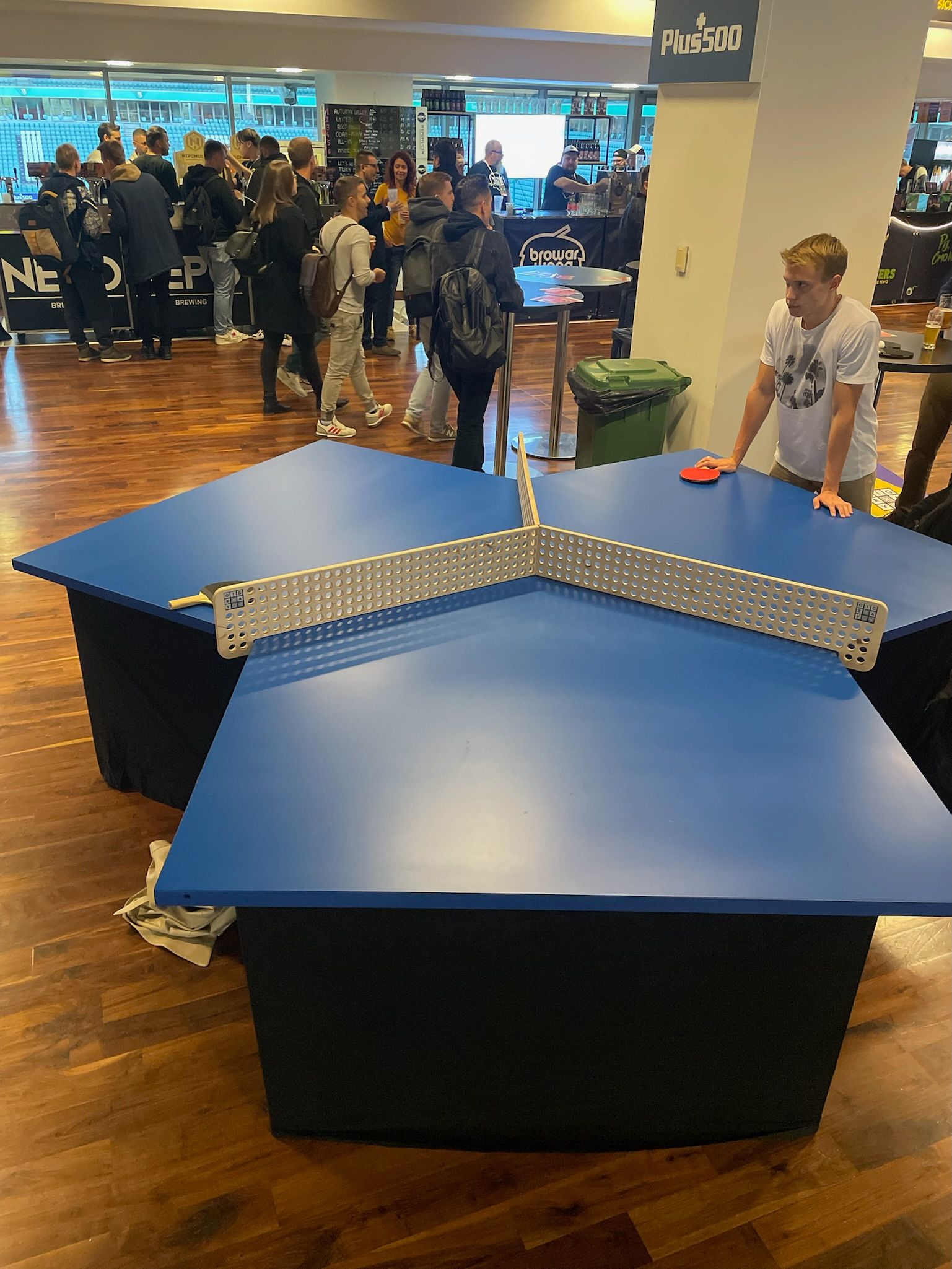 grupa ludzi stoi wokół niebieskiego stołu do ping-ponga w kształcie litery Y.