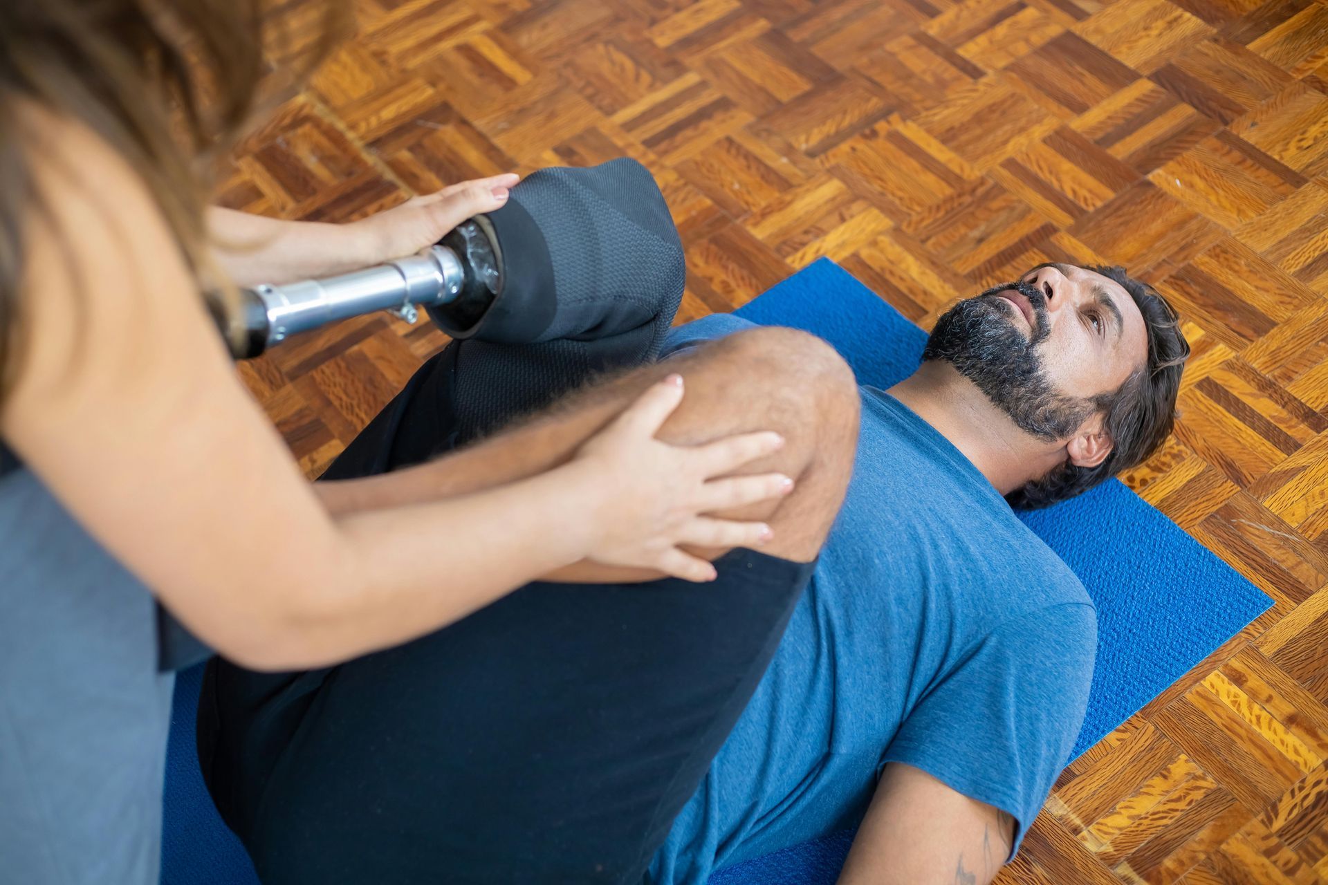 A man is laying on a yoga mat while a woman holds a dumbbell over his knee.