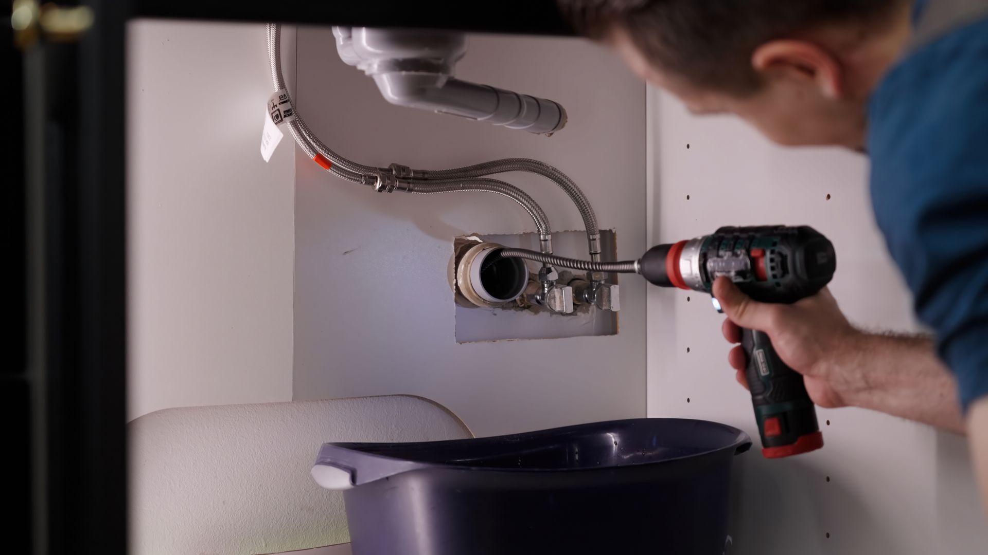 Person using a drill to work on a sink's plumbing inside a cabinet, with a bucket below.