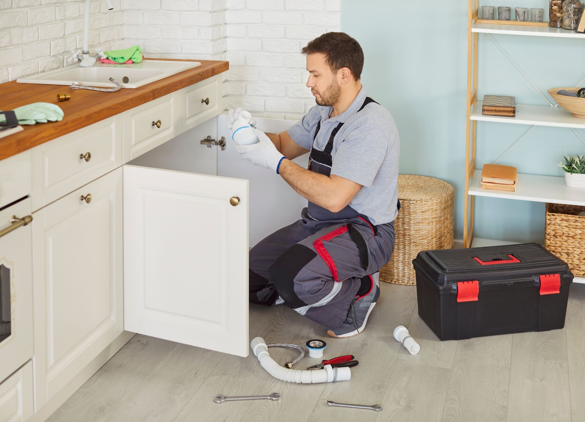 A plumber in a uniform with a toolbox sitting near the sink, fixing a broken pipe in a kitchen.