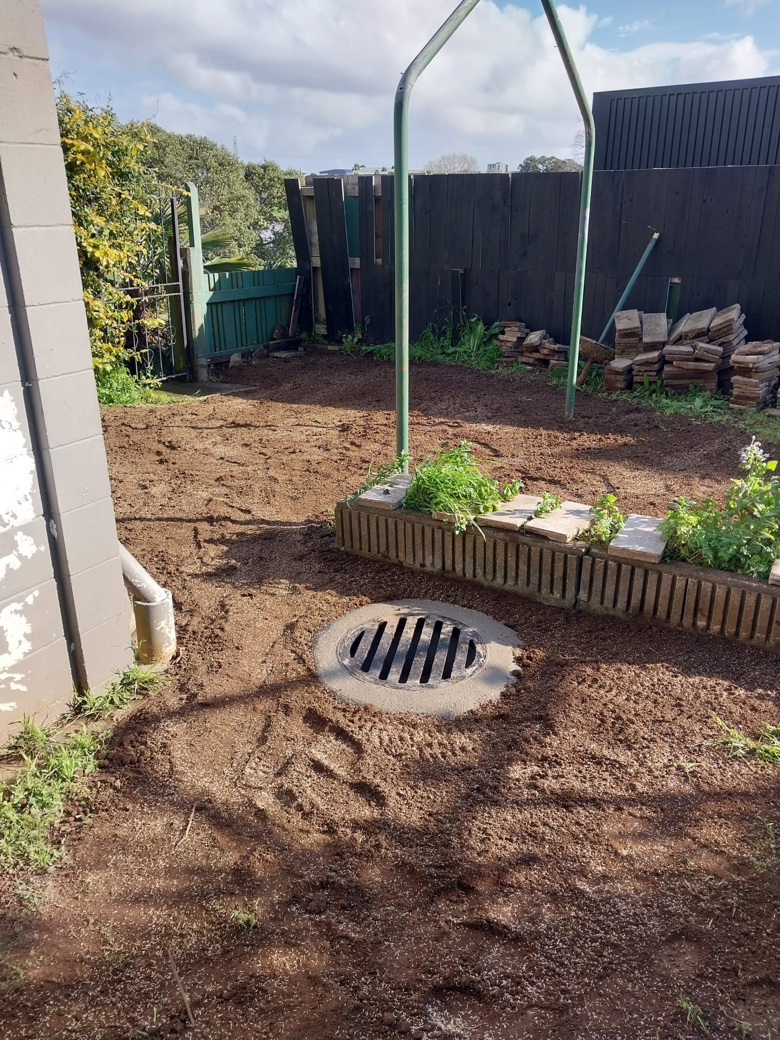 Backyard scene with drain, soil, and garden bed against a fence.