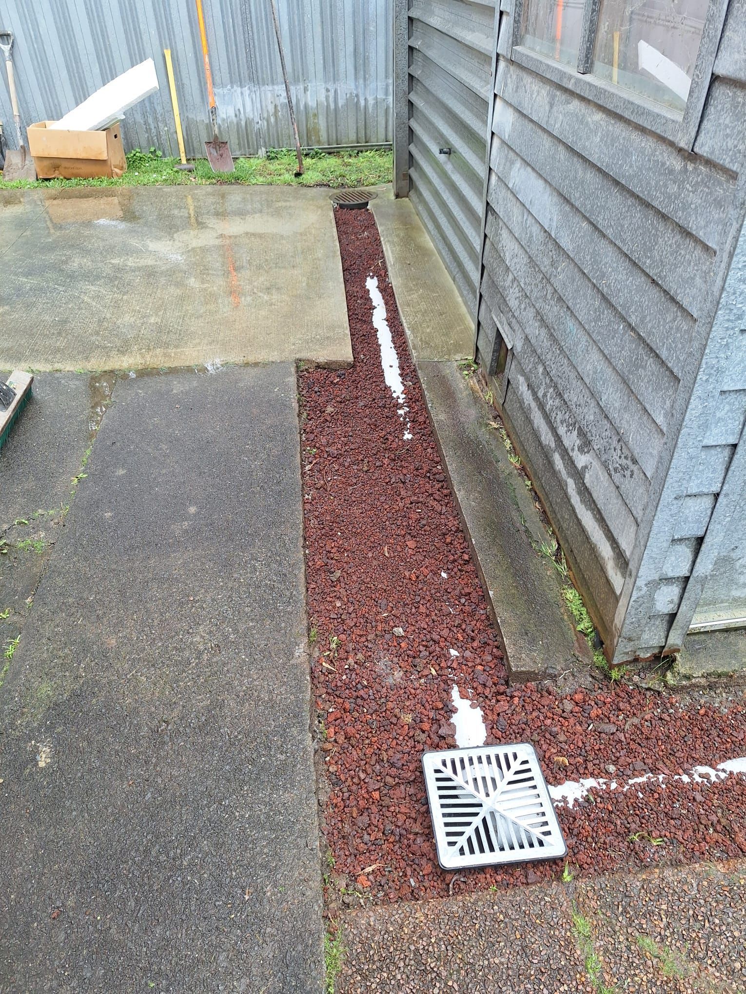 Red rock-filled trench with a drain grate near a concrete patio and a gray building.
